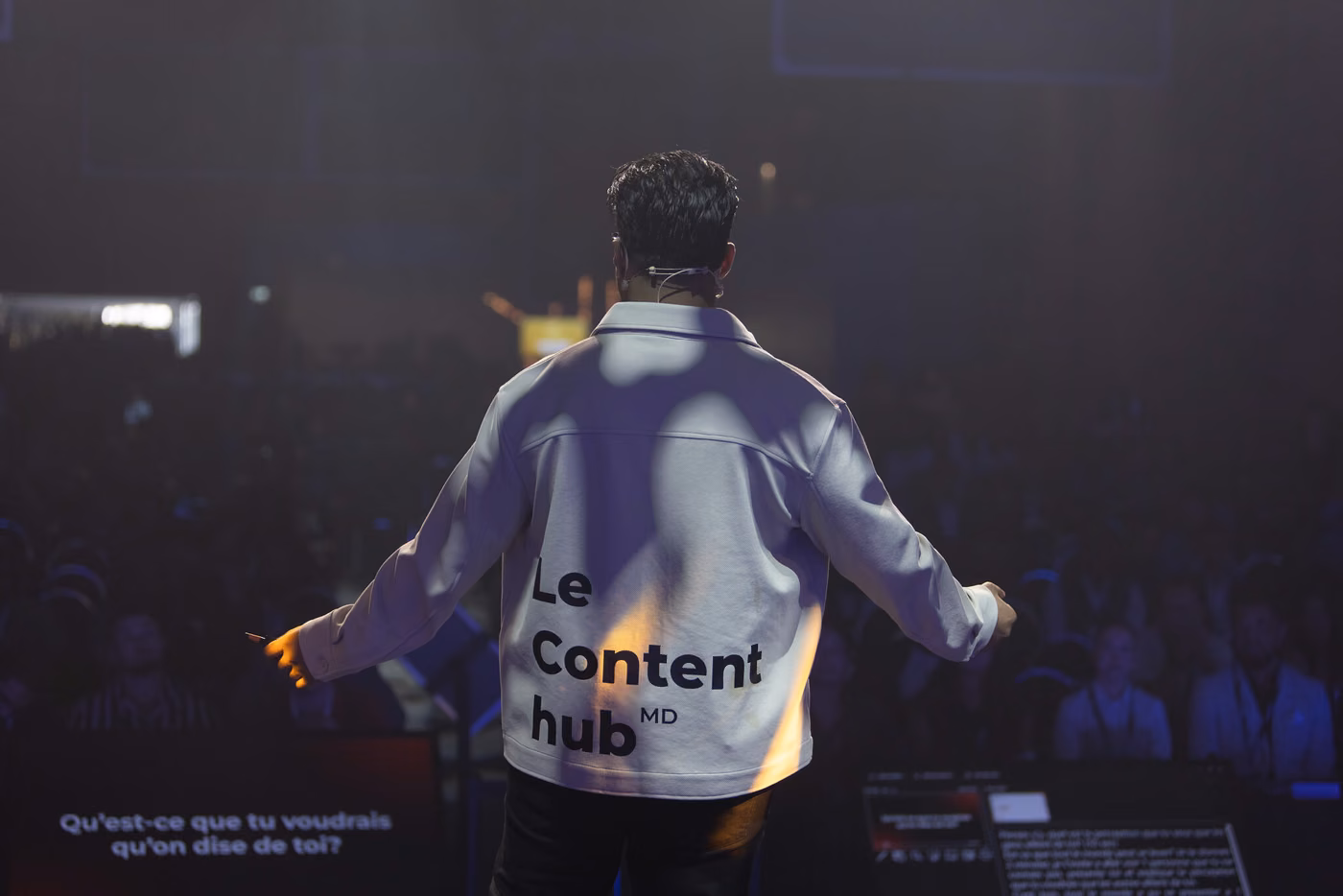 Gwenaël Mercier de dos sur scène, bras ouverts vers une salle comble, devant une diapositive demandant ce qu'on voudrait qu'on dise de soi