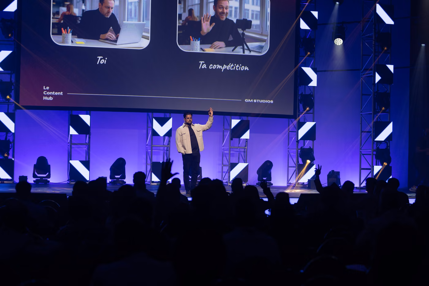 Le conférencier bras levé sur scène, devant un écran géant comparant deux vidéos côte à côte, « Toi » et « Ta compétition »
