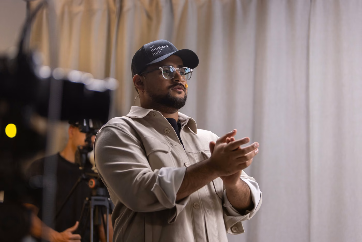 Gwenaël Mercier en casquette et veste beige, mains jointes, en tournage devant un rideau clair et une caméra sur pied