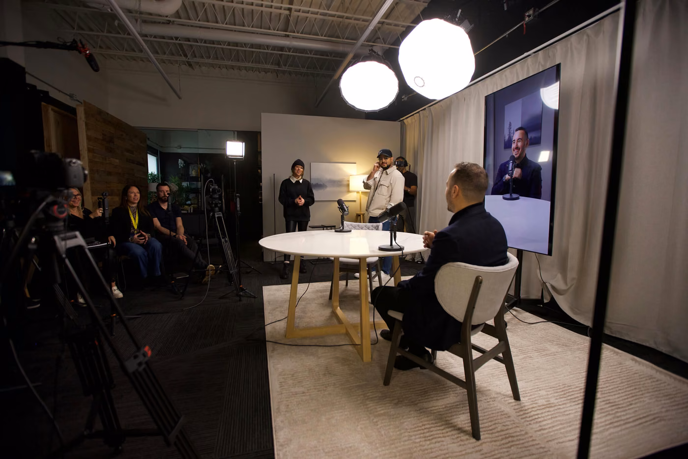 Tournage d'une entrevue en studio : deux personnes assises de part et d'autre d'une table ronde, écran de retour, caméras et public autour